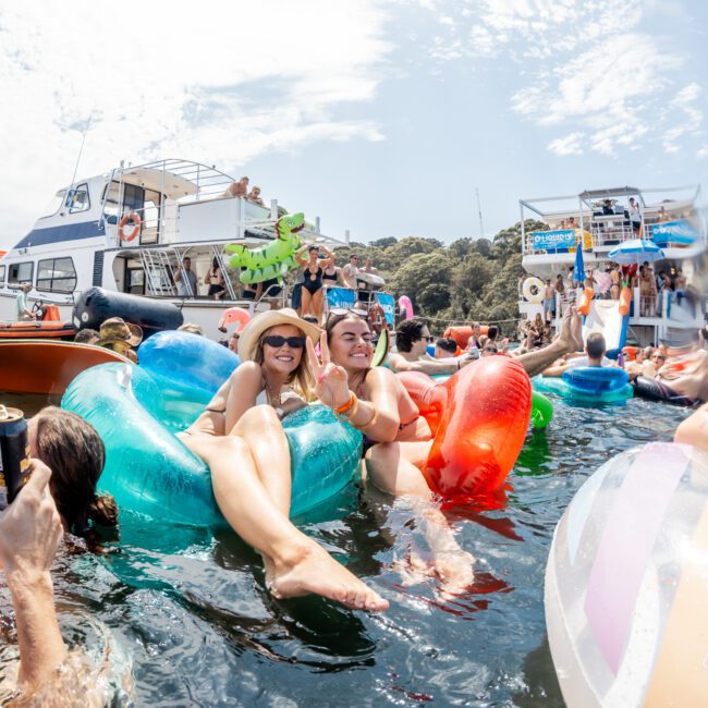 People relax on colorful inflatables in the water, holding drinks and smiling. Boats and more people are in the background, with trees and a bright sky above, creating a lively party atmosphere.
