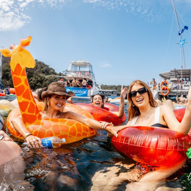 A group of people smiling and relaxing in the water on colorful pool floats, including a giraffe and a chili pepper, with boats and a blue sky in the background.