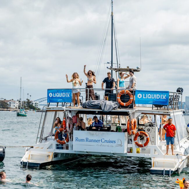 A double-decker boat labeled “Rum Runner Cruises” on the water, with people dancing and relaxing on the upper and lower decks, while others swim nearby under a cloudy sky.