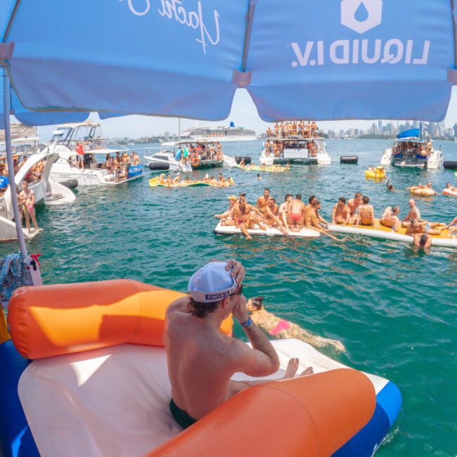 A man sits under a blue umbrella on an inflatable chair, overlooking a crowded water party with people on boats, paddleboards, and floats in a bay. The city skyline is visible in the background.