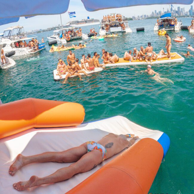 A man in a swimsuit lies face down on an orange inflatable raft in the water, surrounded by boats and groups of people socializing and sunbathing at a lively boat party near a city skyline.