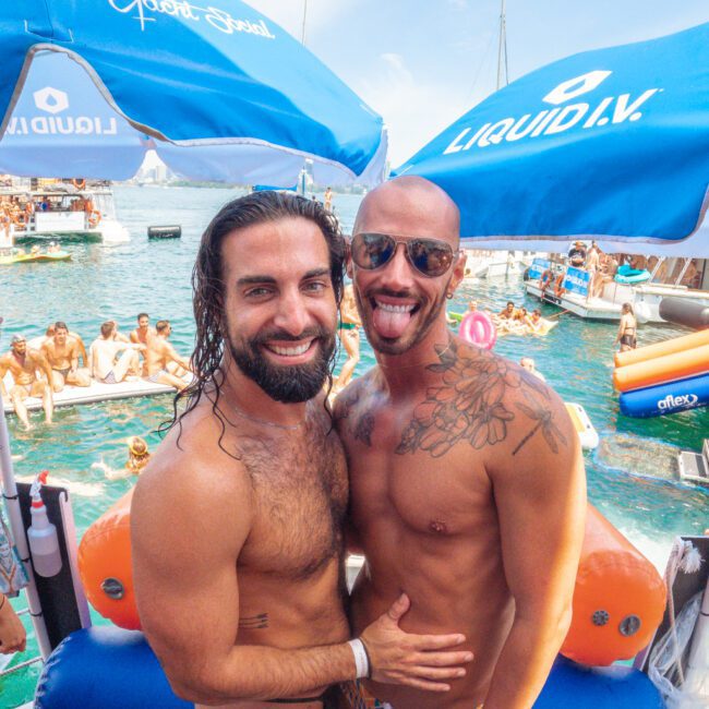 Two men in swimwear smile and pose together at a lively pool party on a dock, with boats, people, and umbrellas in the background. The scene is sunny and festive by the water.