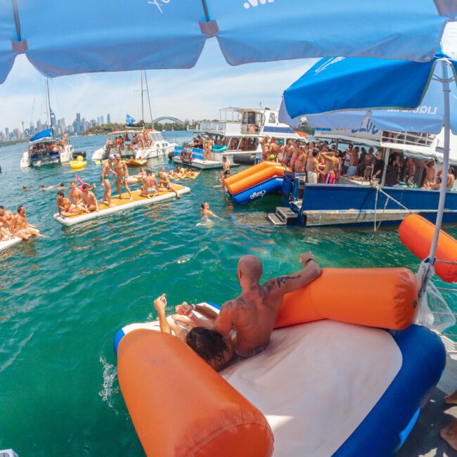 A lively pool party on the water with people lounging on inflatable rafts and swimming near boats. The scene is festive, with groups enjoying the sun, and a large blue and white umbrella is visible in the foreground.