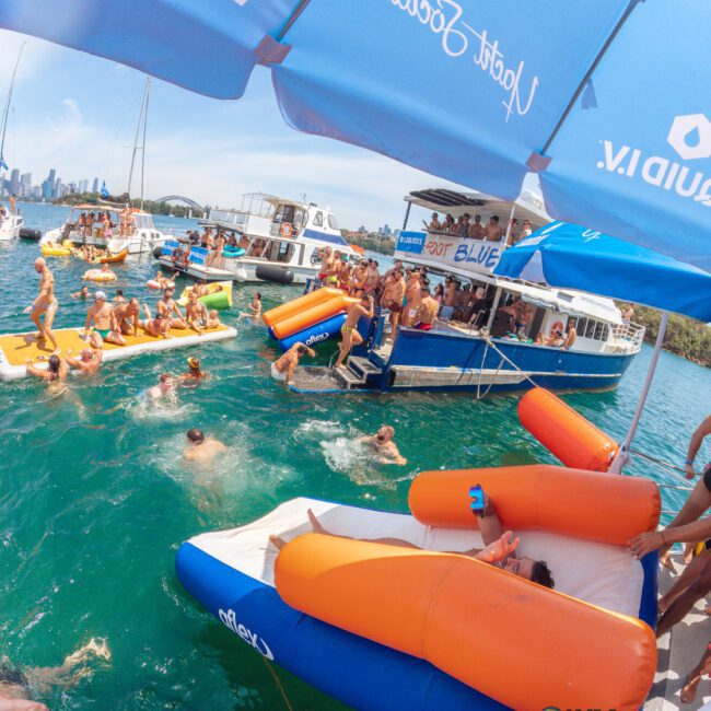 People enjoy a lively boat party on turquoise water, swimming and lounging on orange floats. Several boats are docked together, and a city skyline is visible in the background under blue skies and umbrellas.