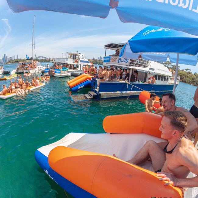 People in swimwear enjoy a sunny day on boats and inflatables in clear blue water. Some are gathered under umbrellas, while others relax or socialize on boats. Skyline and greenery are visible in the background.