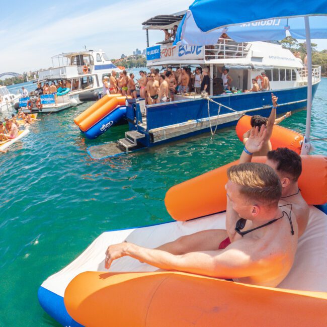 Two men relax on floating orange loungers in clear water near a party boat crowded with people under umbrellas, with other boats and swimmers nearby on a sunny day.