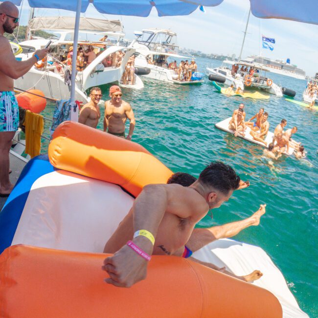 A group of people in swimsuits enjoy a sunny day on boats and floating platforms in blue-green water. Two men are about to slide into the water from a large inflatable on a boat, while others relax and socialize nearby.