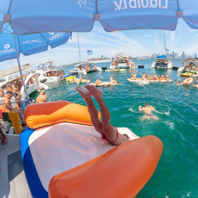 A person in swim trunks slides headfirst down an inflatable water slide from a boat into the ocean, surrounded by other boats, people swimming, and a city skyline in the background.