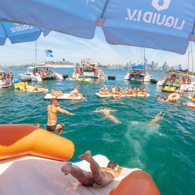 People enjoy a lively boat party on a sunny day; a man slides off an orange inflatable into the water while others swim, lounge on floats, and relax on boats under a blue sky with city buildings in the background.