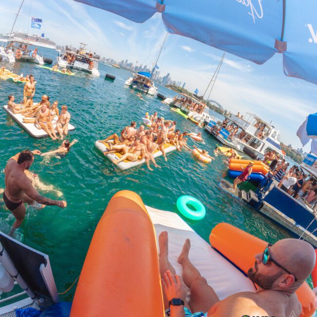 People enjoy a lively pool party on floating inflatables and boats in a sunny, blue lake with a city skyline in the background. A man relaxes on an orange float in the foreground under a branded umbrella.