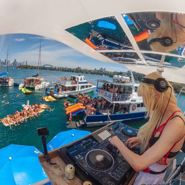 A DJ plays music on a boat deck for a lively crowd on nearby boats and floating rafts, with people enjoying the water under a sunny sky. City skyline is visible in the background.