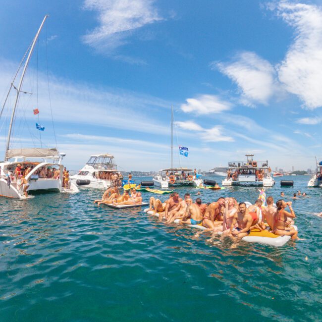 A group of people relax on floating mats in clear blue water, surrounded by several boats under a sunny sky. The scene is lively, cheerful, and festive, with blue flags visible on some boats.