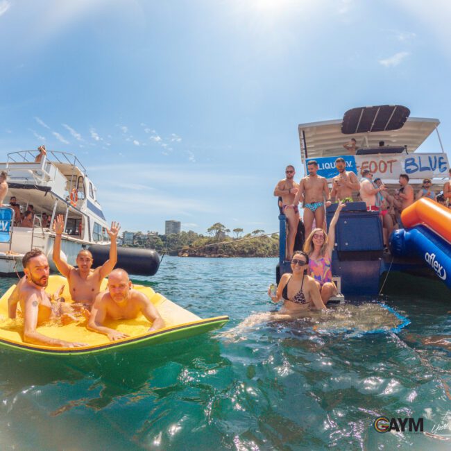 A group of people enjoy a sunny day on the water, swimming, lounging on floats, and gathering on boats. Some wave to the camera, while others relax or stand on a floating platform with a slide. The mood is festive and joyful.