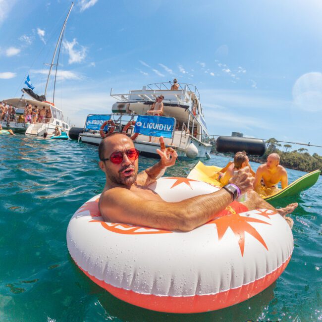 A man wearing red sunglasses lounges in a swimming pool float surrounded by people on boats and paddleboards, enjoying a sunny day on the water. The scene is festive, with clear blue skies and vibrant activity.