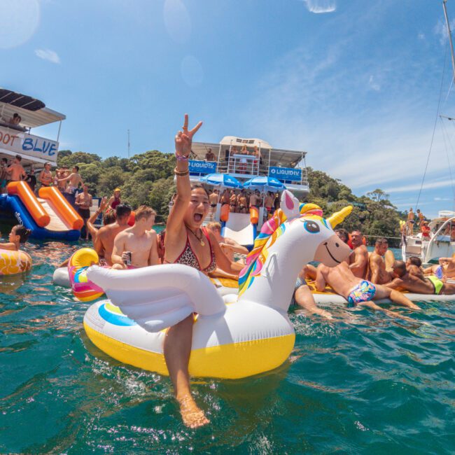 A woman in a bikini sits on a unicorn float, flashing a peace sign among a lively crowd of people swimming and partying near boats on a sunny day.