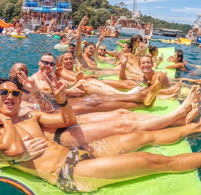 A group of smiling people in swimsuits relax on a floating mat in clear blue water at a lively boat party, with other boats and partygoers in the background under a sunny sky.