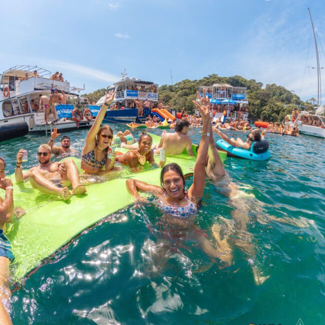 A group of people in swimsuits smile and wave while lounging on a green float in bright blue water, surrounded by boats and other partygoers under a sunny sky.