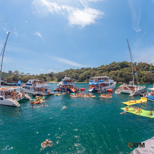 Boats and yachts are gathered on a sunny day in blue-green water, with many people swimming, lounging on inflatables, and socializing. Lush green trees and a blue sky are in the background.