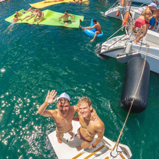 Two smiling men in swimwear stand on the back deck of a boat, waving at the camera. Behind them, people relax on large floaties in clear blue water, and several boats are anchored close together for a lively party.