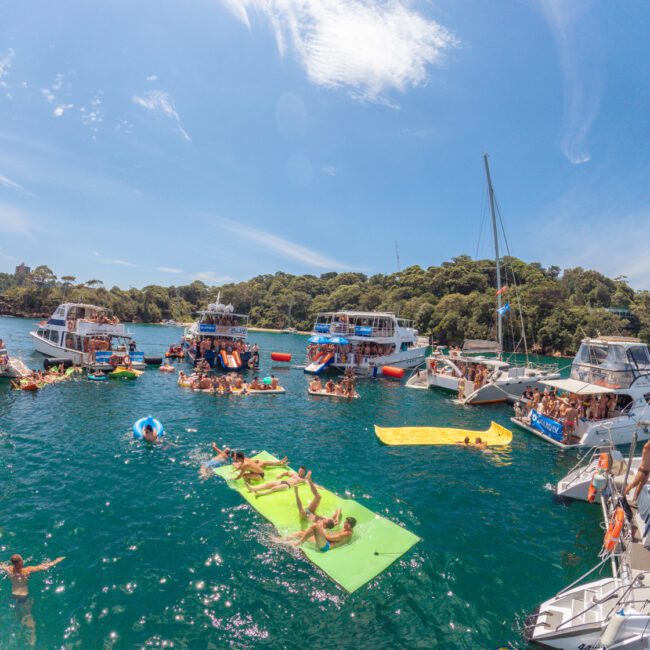 A lively scene of people enjoying a boat party on a sunny day, with several boats anchored close together, people swimming, and others relaxing on floating mats in clear blue water surrounded by trees.