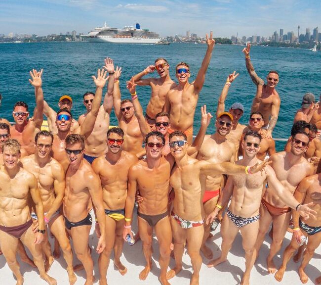 A group of men in swimwear cheerfully pose on a boat under a blue sky, with city skyscrapers and a cruise ship visible in the background. The water is a bright turquoise, and everyone looks happy.