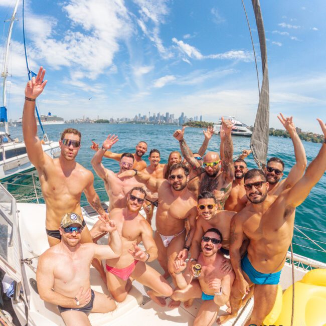 A group of smiling men in swimsuits pose and wave enthusiastically on a sailboat, with blue skies, water, and a city skyline in the background.