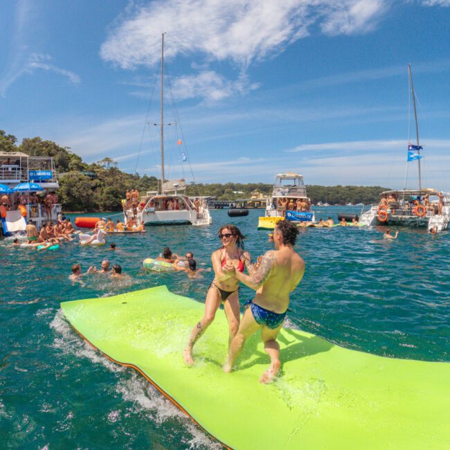 Two people laugh and hold hands on a floating mat in blue water, surrounded by boats and other people enjoying a sunny day, with trees and a blue sky in the background.
