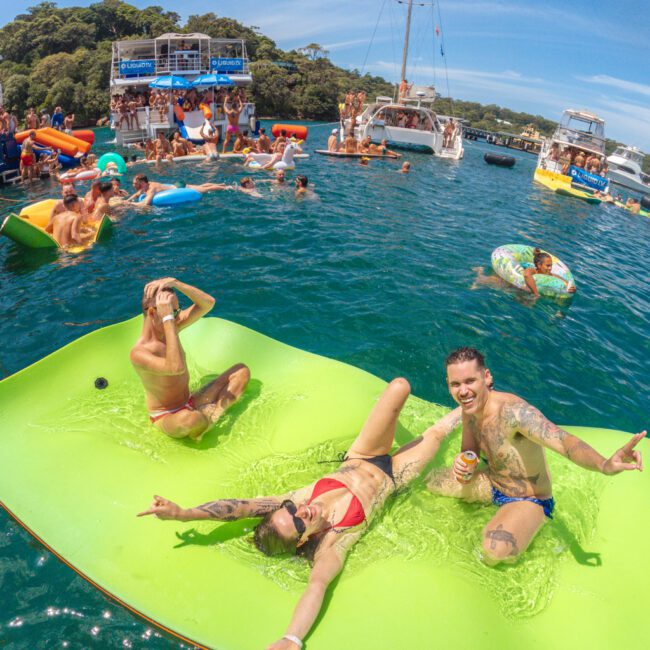 People relax on a large green float in the water, smiling and enjoying drinks. In the background, more people swim, float, and gather on boats at a lively party under a sunny sky. Logo "GAYM" appears in the corner.