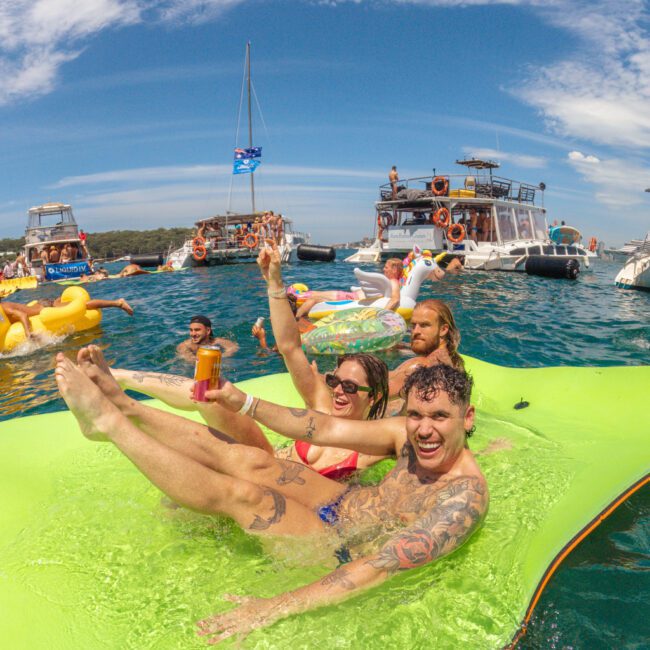 Three people laugh and raise drinks while relaxing on a green float in the water, surrounded by boats and other people on inflatables, under a partly cloudy blue sky.