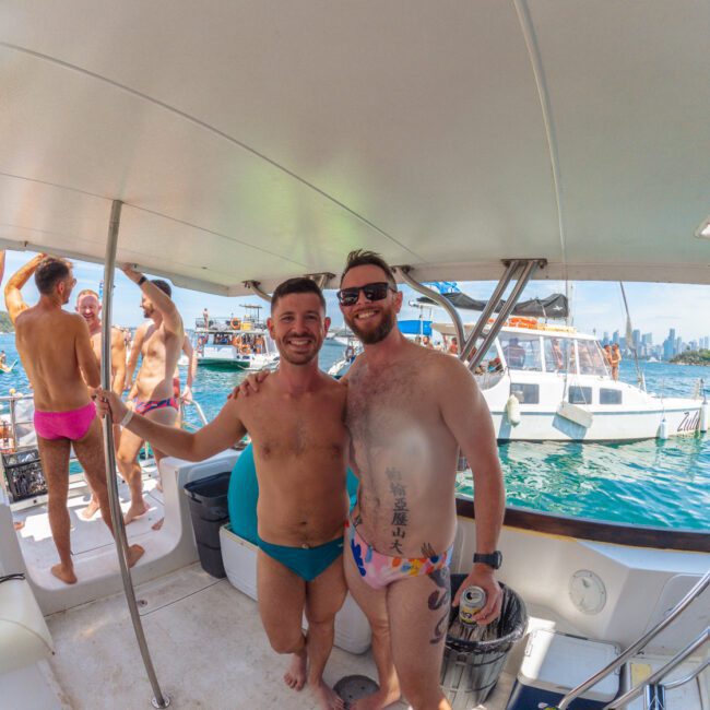 Two smiling men in swimsuits stand arm-in-arm on a boat deck at a lively party near the water. Other people in colorful swimwear and boats are visible in the background under a sunny sky.