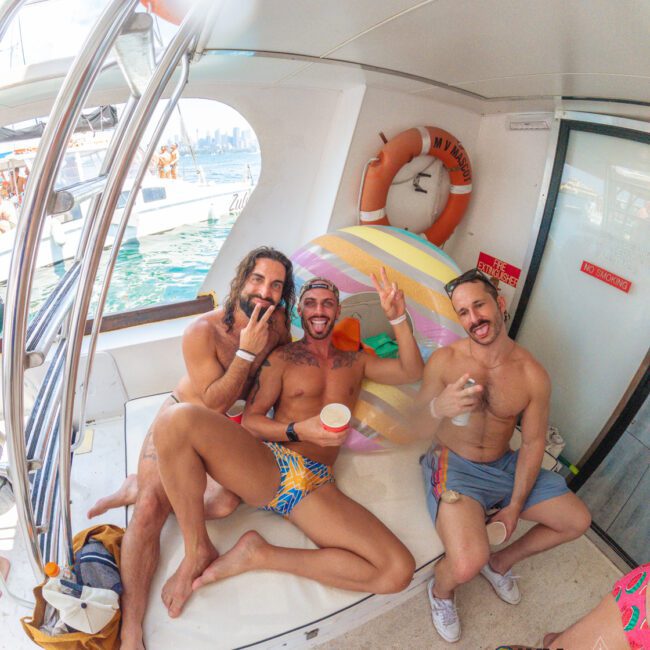 Three men in colorful swimwear sit on a boat, smiling and making peace signs. A rainbow float and life preserver are in the background. They look happy and relaxed, enjoying a sunny day on the water.