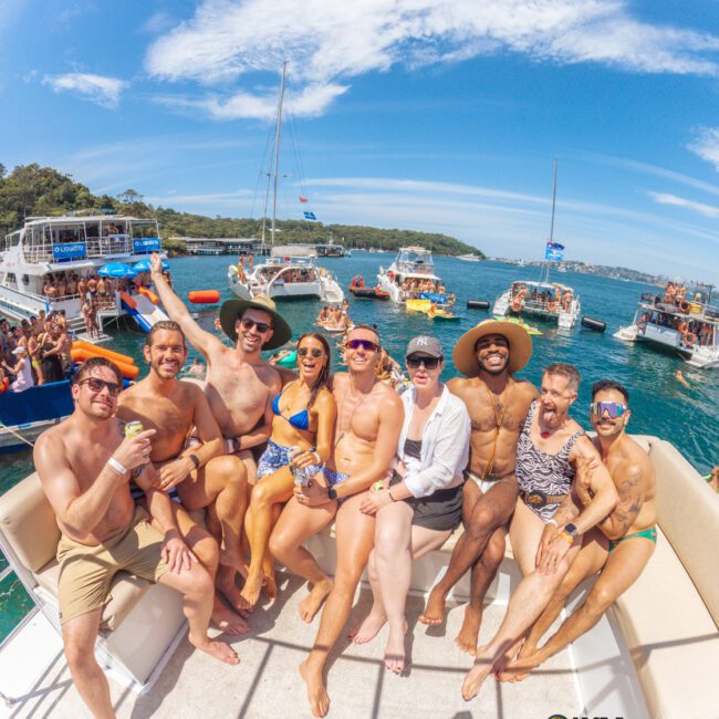 A group of eight people in swimwear smile and pose together on a boat, with other party boats and people in the background on a sunny day, surrounded by blue water and clear skies.