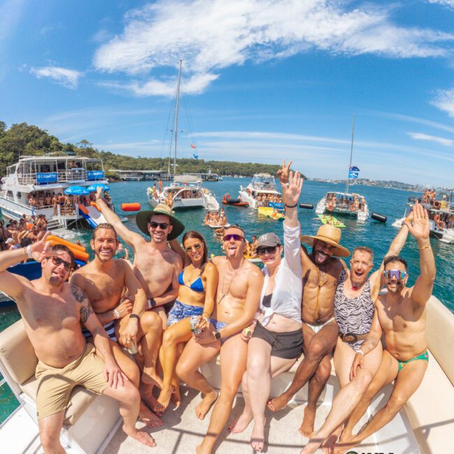 A group of people in swimwear sit and pose cheerfully on a boat, surrounded by other boats on a sunny, blue-sky day. The atmosphere is festive, with clear water and a crowd enjoying the event.