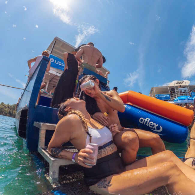 Two people relax on a boat’s swim platform; one sits holding a drink and laughing, while the other adjusts swim goggles. Other people are on the boat, and bright sunlight shines overhead. The water and inflatable floats are visible nearby.