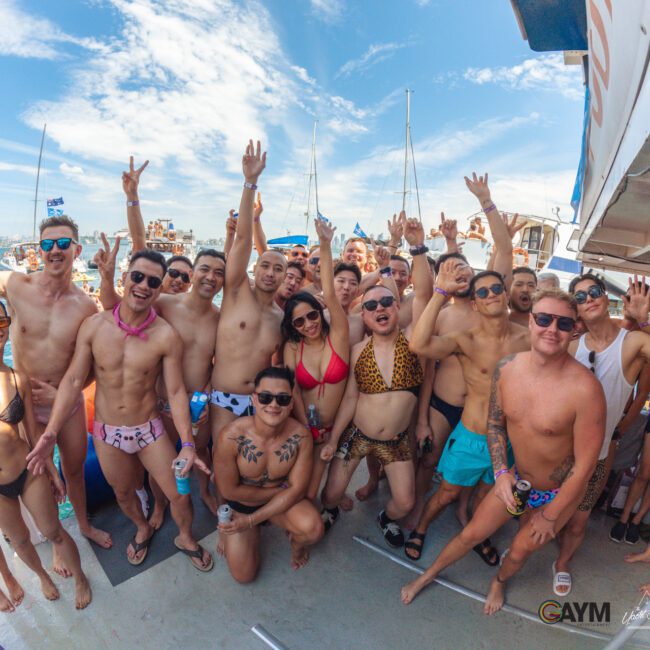 A large group of people in swimsuits pose and smile on a boat under a blue sky, some raising their arms in celebration. Other boats and water are visible in the background.