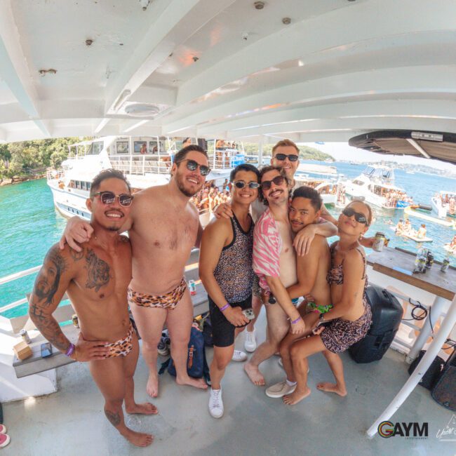 A group of people wearing animal print swimwear pose and smile together on a boat, with bright sunlight, blue water, and other boats and partygoers in the background. The scene is festive and lively.