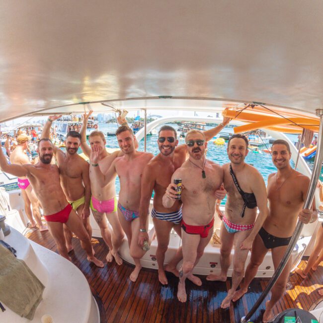A group of men in colorful swimwear smiling and posing together on a boat, with the ocean and other boats visible in the background under sunny weather.
