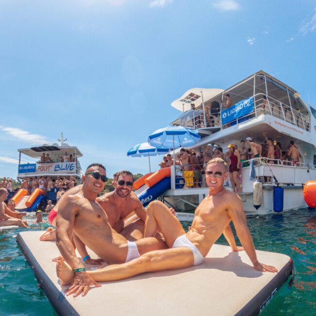 Three men in white swim trunks pose and smile on a floating mat in the sea, with party boats and a crowd of people in swimwear enjoying the water and sunshine in the background.
