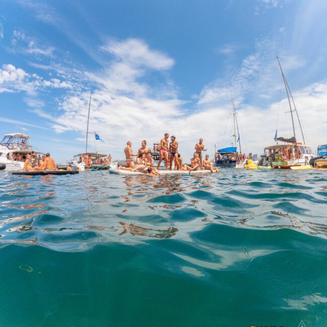 A group of people on floating platforms and boats enjoy a sunny day on the water, surrounded by yachts and sailboats under a partly cloudy blue sky.