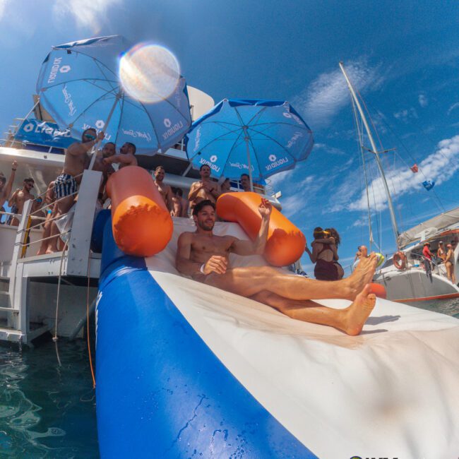 A man slides down an inflatable slide into the ocean from a boat, surrounded by people enjoying the sun, with blue umbrellas and a sailboat in the background under a clear sky.