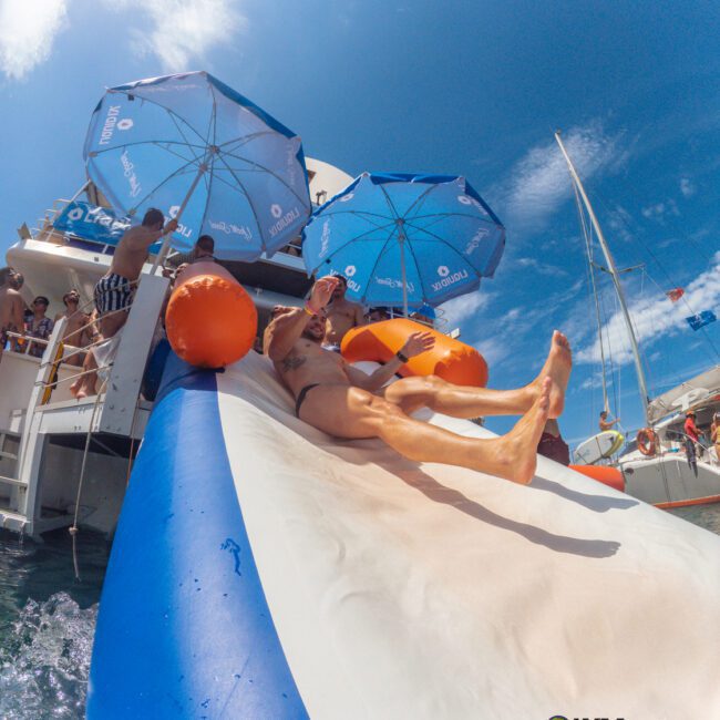 A person slides down an inflatable water slide from a boat into the sea, with blue umbrellas and people watching under a bright, sunny sky.