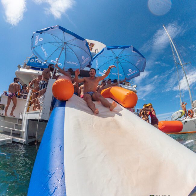 A smiling man slides down an inflatable water slide from a boat into the sea, surrounded by people on the boat under blue umbrellas, with a sunny sky and sailboats in the background.