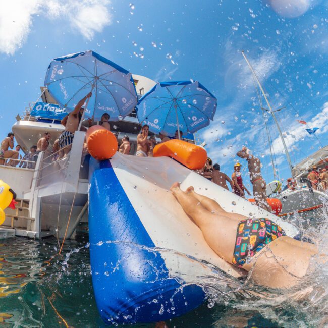 A person slides down a blue and white inflatable water slide into the sea from a boat, surrounded by people, blue umbrellas, and a sunny sky.