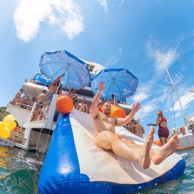 A man slides down an inflatable slide from a boat into the water, arms raised and smiling, while people watch and relax under blue umbrellas on the boat deck under a clear blue sky.