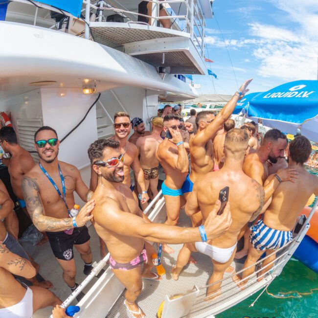 A large group of men in swimsuits are partying on a boat in sunny weather, smiling, dancing, and taking selfies. A blue tent with "Liquid Sky" is visible, and the vibe is lively and festive.