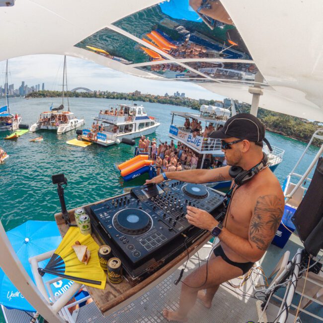 A DJ in swimwear mixes music on a boat deck, overlooking a lively crowd on nearby boats in a sunny, waterside party scene. The water and city skyline are visible in the background.