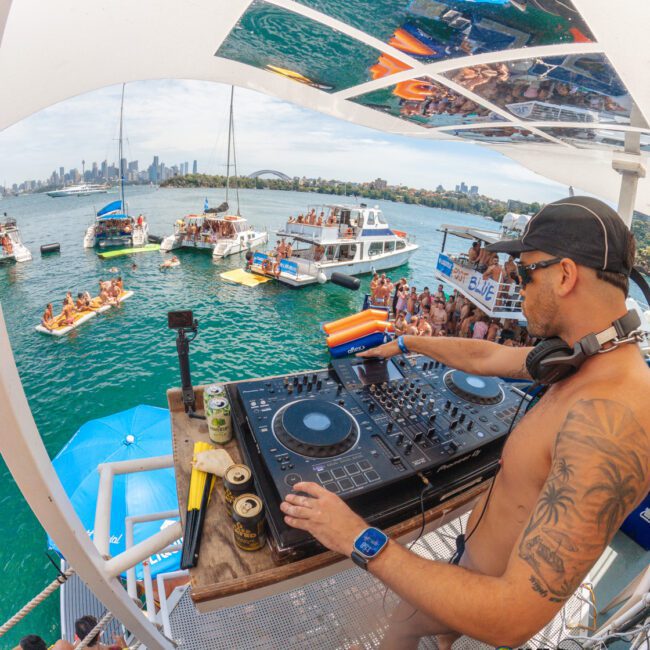 A shirtless DJ wearing sunglasses and a cap plays music on a boat deck, overlooking a crowd of people partying on nearby boats and floating platforms on blue water, with a city skyline in the background.
