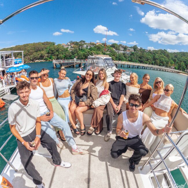 A group of young adults sit and stand on a boat deck, smiling at the camera on a sunny day, with a marina, clear blue sky, and green trees visible in the background.