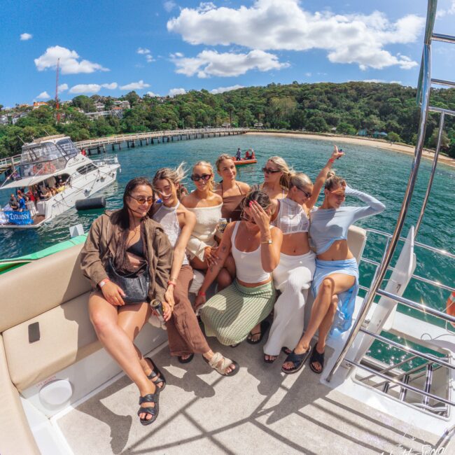 A group of eight women in summer outfits sit and pose for a photo on a boat, smiling and enjoying a sunny day with clear blue water and a scenic, green coastline in the background.