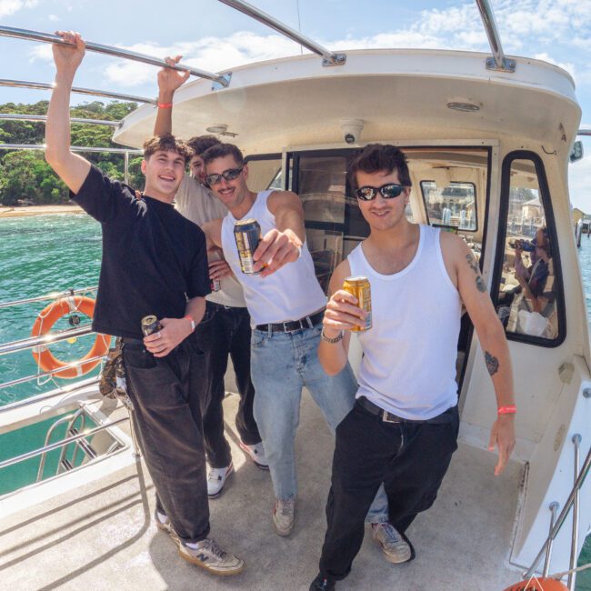 Four young men smiling and holding drinks stand on the deck of a boat under a sunny sky, with water and greenery visible in the background. They appear to be enjoying a casual day out on the water.